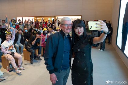 Tim Cook, CEO of Apple, taking a selfie with a woman holding a yellow iPhone, surrounded by a large crowd of people inside an Apple Store.