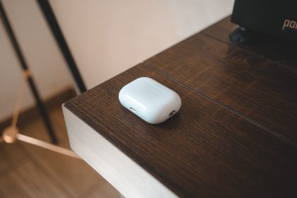 ALT Text: The image shows a white AirPods Pro 2 case placed on a wooden surface. The case is sleek and compact, with a smooth glossy finish. It has an LED indicator on the front and a charging port at the bottom. The wooden surface contrasts with the modern look of the AirPods case, highlighting its design. The background includes parts of furniture and a tripod-like structure, adding a minimalist and modern aesthetic to the setting.