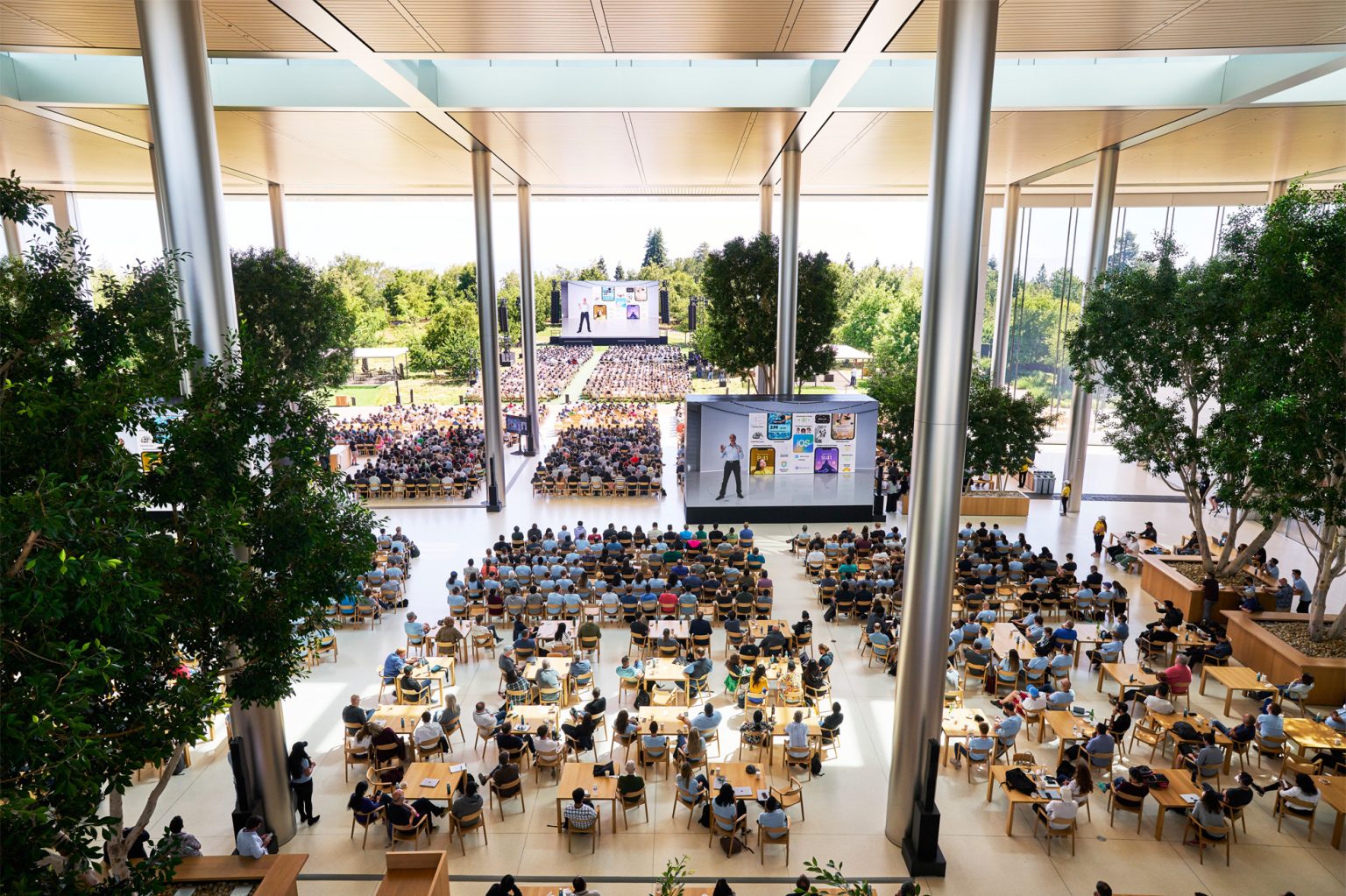 The image shows a large gathering at Apple Park for the WWDC22 keynote presentation. The audience is seated both indoors and outdoors, with the main stage visible in the background displaying an Apple presenter and various slides about iOS 16. The interior space is modern and spacious, featuring tall glass walls, large trees, and a clean, minimalist design with numerous tables and chairs occupied by attendees.