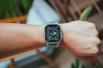 The image shows an Apple Watch Ultra on a person’s wrist. The watch has a rugged design with a gray titanium case and a colorful, detailed watch face displaying various metrics such as activity rings, date, and battery percentage. The watch band is a sport loop in a blue and gray color scheme. The background is slightly blurred, featuring a wooden chair and some greenery, highlighting the watch as the main focus.