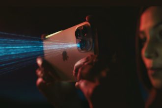 Close-up of a woman holding an Apple iPhone with a LiDAR sensor, showing blue laser beams added in post-production, in a dimly lit environment