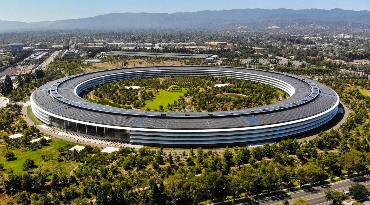 Aerial view of Apple Park, the circular headquarters of Apple Inc. in Cupertino, California, surrounded by greenery and featuring a colorful rainbow structure in the center, with the Silicon Valley landscape in the background