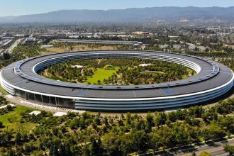 Aerial view of Apple Park, the circular headquarters of Apple Inc. in Cupertino, California, surrounded by greenery and featuring a colorful rainbow structure in the center, with the Silicon Valley landscape in the background