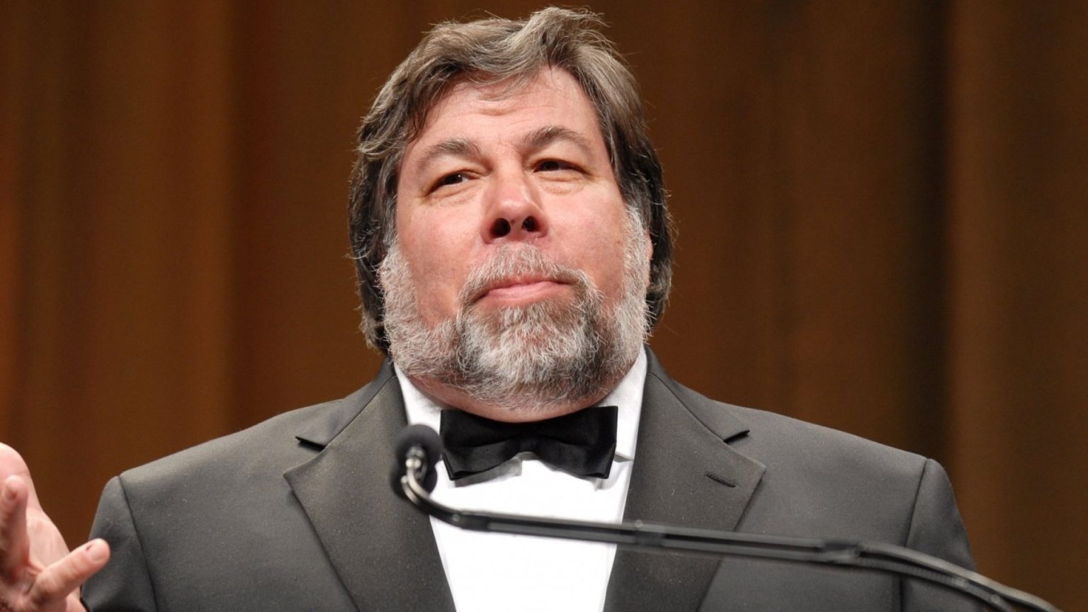 Steve Wozniak, co-founder of Apple Inc., standing at a podium in a black tuxedo with a white shirt and black bow tie, delivering a speech. He has a gray beard and is set against a blurred, warm-colored background