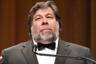 Steve Wozniak, co-founder of Apple Inc., standing at a podium in a black tuxedo with a white shirt and black bow tie, delivering a speech. He has a gray beard and is set against a blurred, warm-colored background