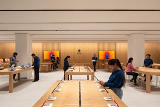 The interior of an Apple Store in Saket, Delhi, India, featuring a spacious and minimalist design with light wooden tables displaying various Apple products like iPads, iPhones, and MacBooks. Several customers and Apple staff, dressed in blue shirts, are engaged with the devices and assisting visitors. The background showcases large digital screens with vibrant, colorful graphics and a central “Pickup” area under the Apple logo.