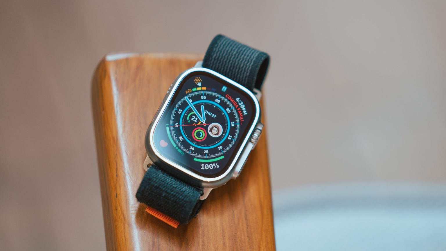 The image shows an Apple Watch Ultra resting on a wooden surface. The watch has a rugged design with a silver titanium case and a black sport loop band. The watch face is illuminated, displaying various metrics such as activity rings, date, air quality index (AQI), and a contact photo