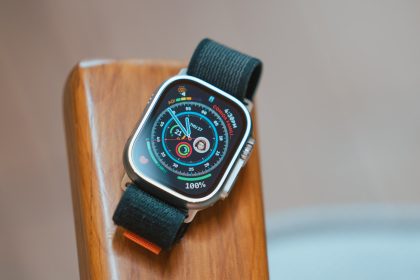 The image shows an Apple Watch Ultra resting on a wooden surface. The watch has a rugged design with a silver titanium case and a black sport loop band. The watch face is illuminated, displaying various metrics such as activity rings, date, air quality index (AQI), and a contact photo