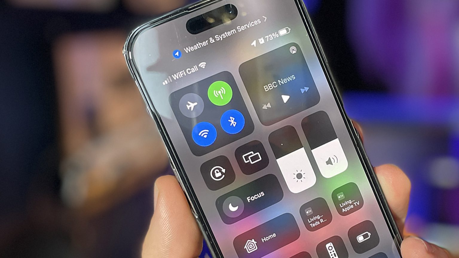 A person holds an iPhone displaying the Control Center on its screen. The Control Center shows various toggles and controls, including WiFi, Bluetooth, airplane mode, and music playback controls. The top of the screen indicates the device is connected to WiFi and shows a battery level of 73%. The background is slightly blurred, focusing attention on the iPhone’s screen.