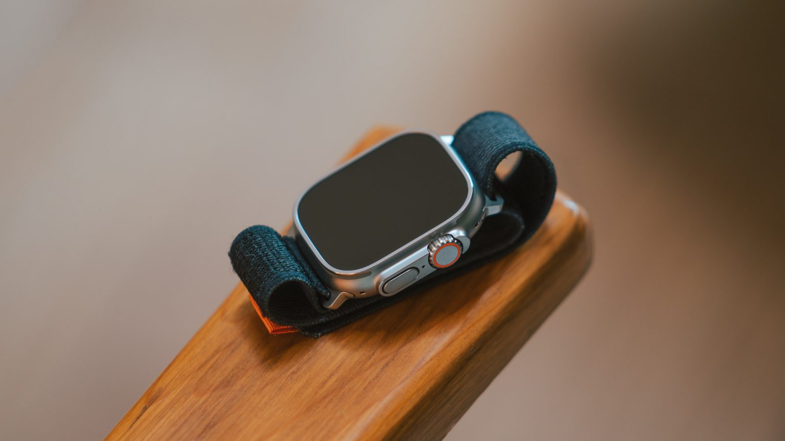 The image features an Apple Watch Ultra with a black band resting on a wooden surface. The watch face is turned off, showcasing the sleek design of the device. The digital crown and side button are visible, highlighting the watch’s robust and premium build quality.