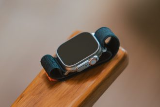 The image features an Apple Watch Ultra with a black band resting on a wooden surface. The watch face is turned off, showcasing the sleek design of the device. The digital crown and side button are visible, highlighting the watch’s robust and premium build quality.