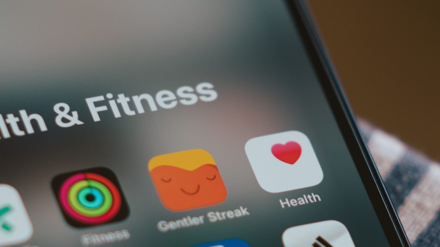 The image shows a close-up of an iPhone screen displaying a folder titled “Health & Fitness.” Within the folder are various app icons, including Apple’s Fitness app with colorful activity rings, the Gentler Streak app with a relaxed face icon, and Apple’s Health app with a pink heart on a white background. The screen is slightly blurred around the edges, focusing on the app icons.