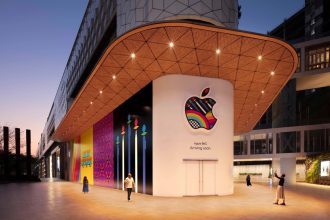 The exterior of the soon-to-be-opened Apple BKC store in India at dusk. The storefront features a colorful, patterned Apple logo and the text “Apple BKC Arriving soon.” The surrounding area includes vibrant, artistic murals and a modern architectural design. People are walking and taking photos outside the store