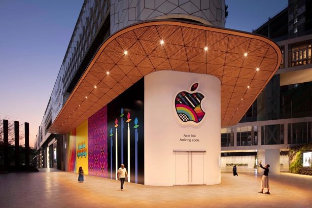 The exterior of the soon-to-be-opened Apple BKC store in India at dusk. The storefront features a colorful, patterned Apple logo and the text “Apple BKC Arriving soon.” The surrounding area includes vibrant, artistic murals and a modern architectural design. People are walking and taking photos outside the store