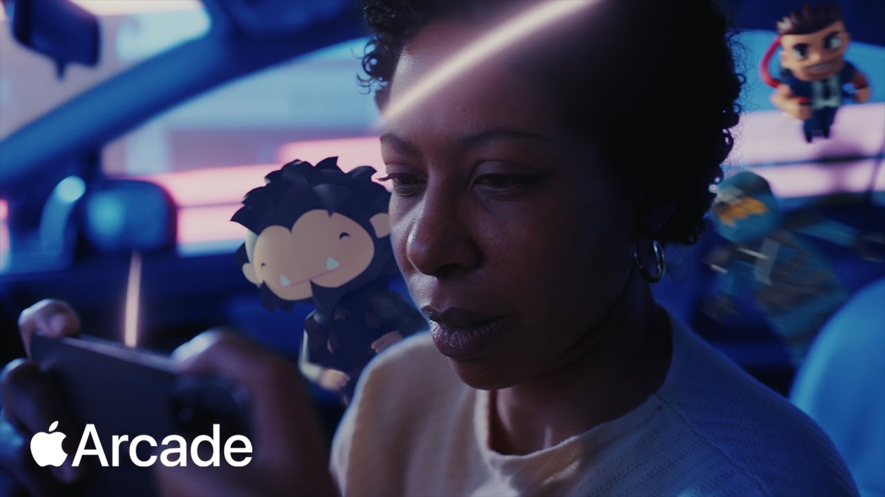 The image is a still from an Apple Arcade advertisement. It features a person intensely focused on playing a game on their mobile device. Animated gaming characters float around them, blending the real and virtual worlds. The background shows the interior of a car with a blurred cityscape outside the windows, giving a sense of immersion in the gaming experience. The Apple Arcade logo is prominently displayed in the bottom left corner.