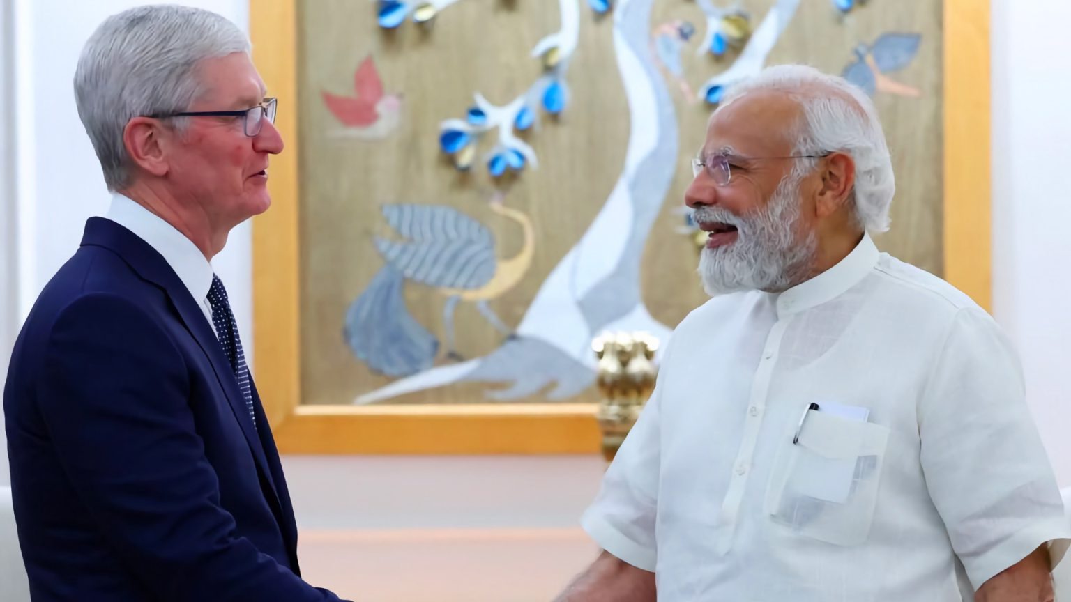 Tim Cook, CEO of Apple, shaking hands with Indian Prime Minister Narendra Modi during a formal meeting. Tim Cook is dressed in a dark blue suit and tie, while Prime Minister Modi is wearing a traditional white kurta. Both are smiling and engaging in a warm handshake, with a colorful decorative background featuring artistic birds and floral designs. The setting suggests a cordial and significant discussion between the two leaders.