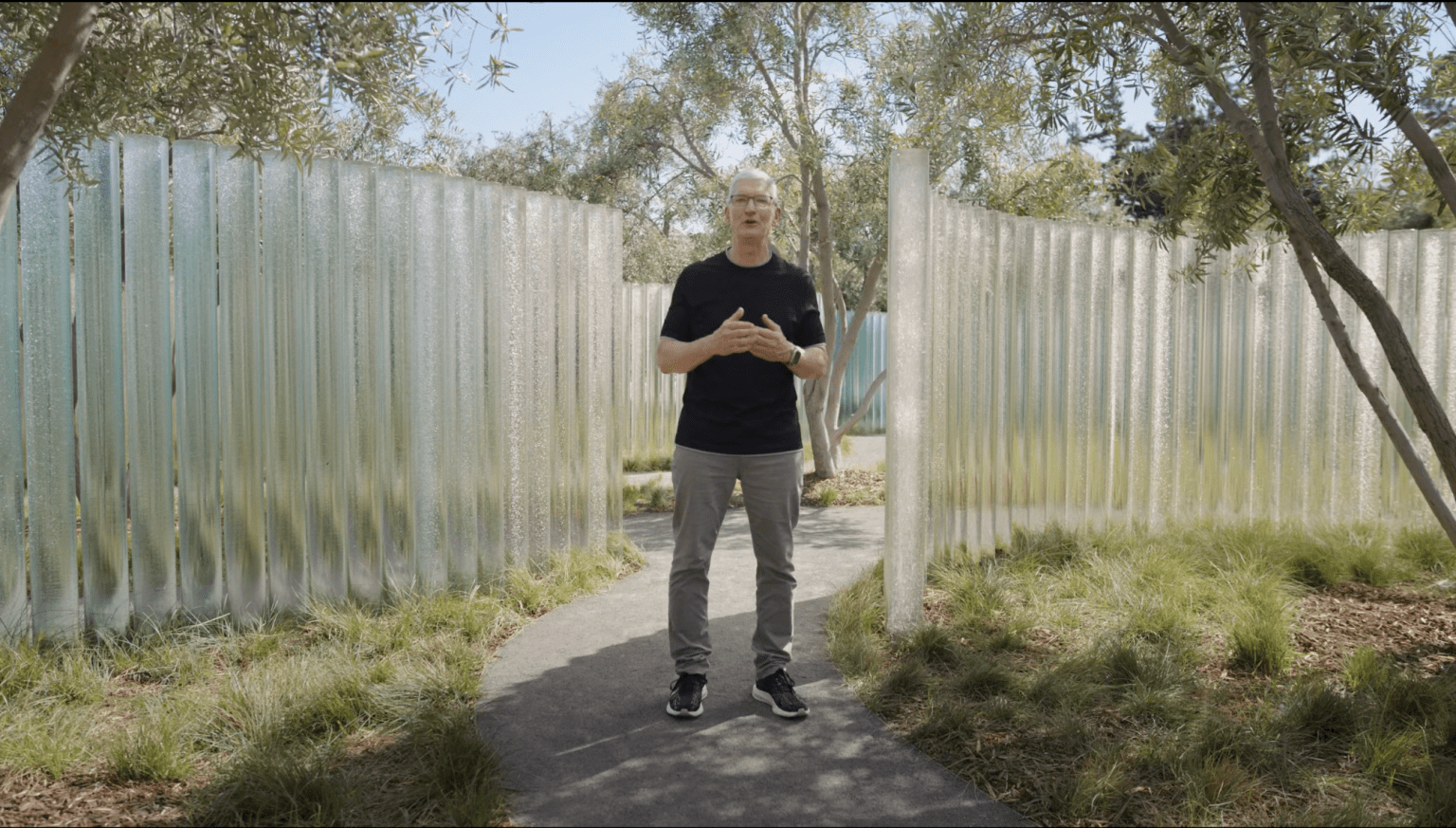 Apple CEO Tim Cook standing outdoors among tall glass pillars and trees, delivering a presentation in a tranquil garden setting